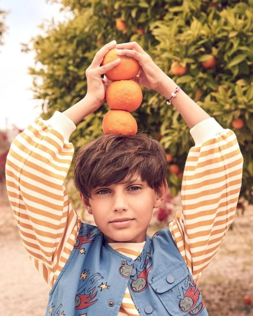 Young boy balances a stack of three oranges on his head in an outdoor orchard, wearing a striped shirt and patterned jacket. 