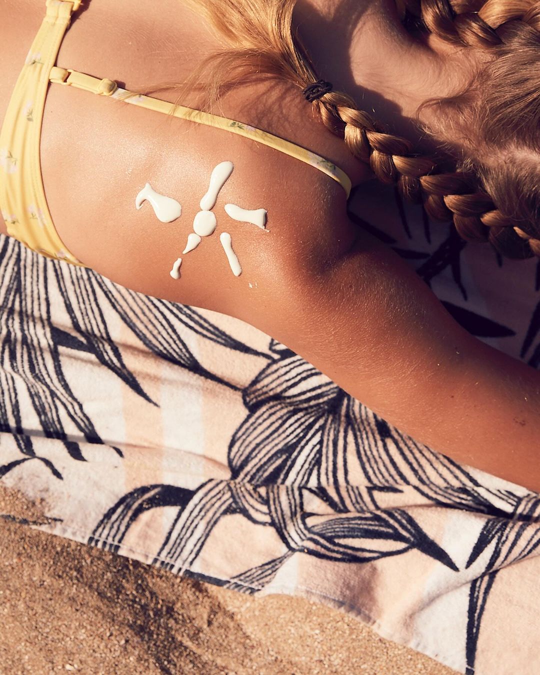Woman on beach lying on towel with sunscreen in sun shape on skin, wearing a yellow bikini top and braided hair.