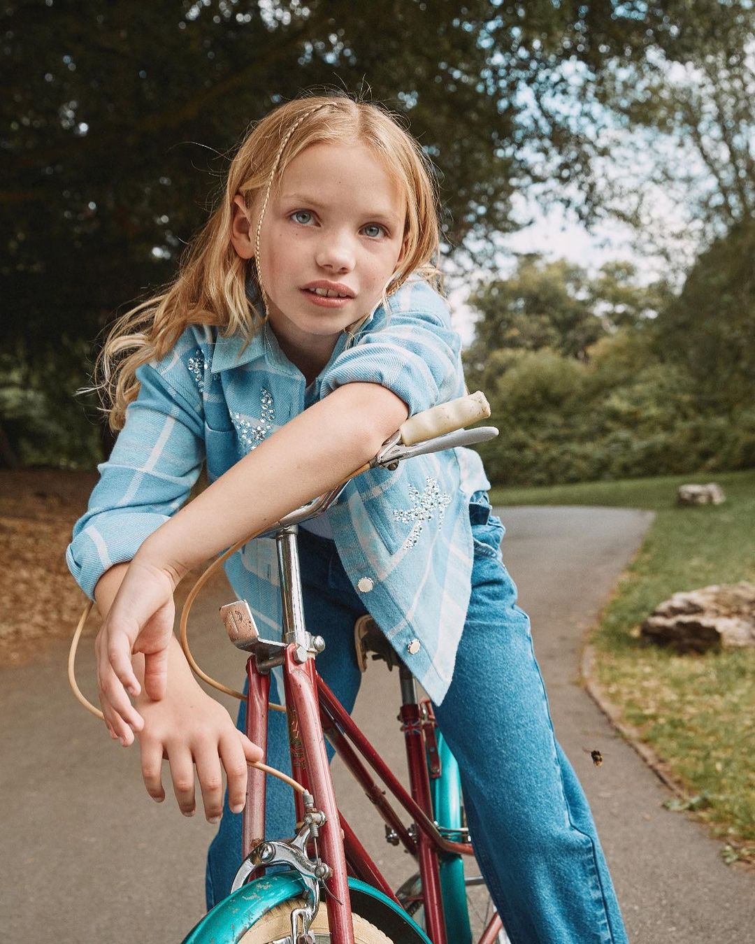 Girl in blue jean outfit rests on the handle bars of an old bicycle with a path and trees in the background in natural lighting.