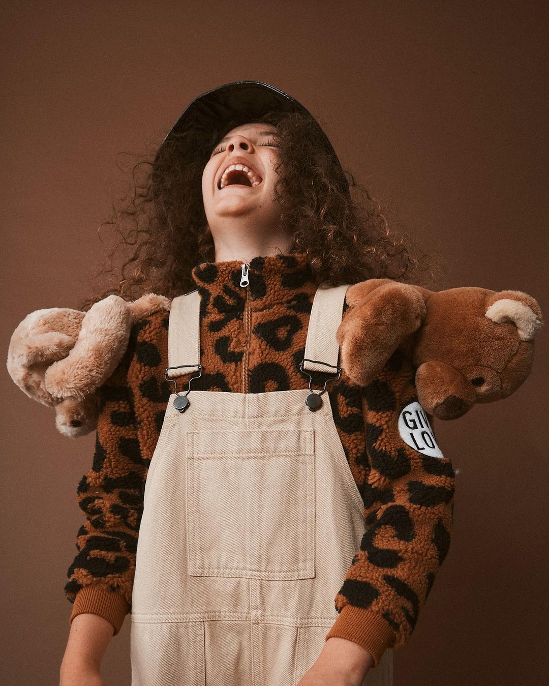 Laughing girl with afro wearing a brown animal print jacket, denim overalls, and a black hat with teddy bears on shoulders.
