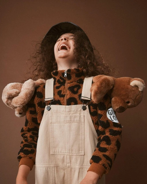 Laughing girl with afro wearing a brown animal print jacket, denim overalls, and a black hat with teddy bears on shoulders.