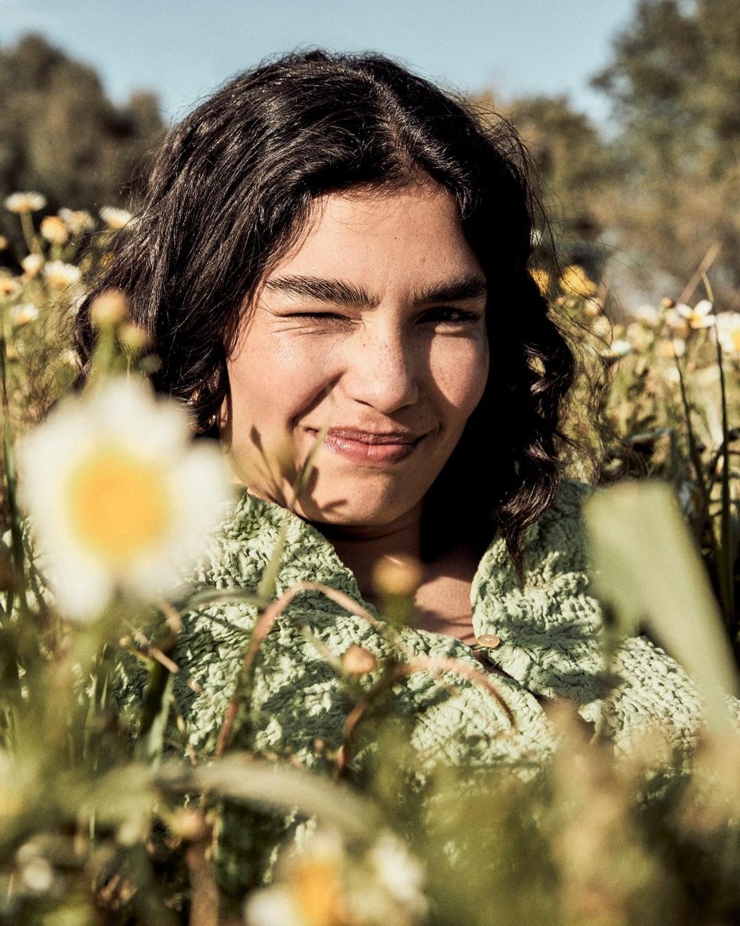 Woman winking in outdoor portrait among field of white and yellow wildflowers wearing a knit green sweater in natural light.