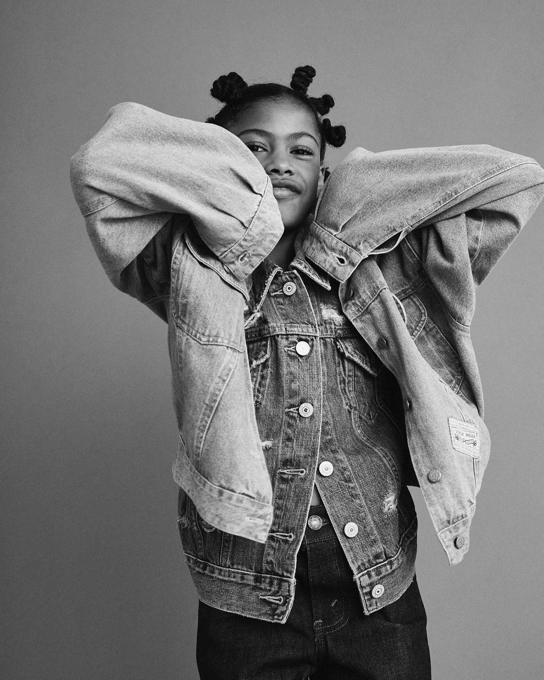 Young child with hair in small knots wearing a denim jacket and dark pants against a plain studio background.