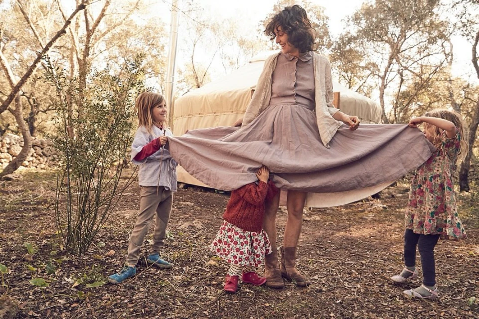 Woman in a dress with two children holding up the skirt, and another child underneath, outdoors in a natural setting near a yurt.