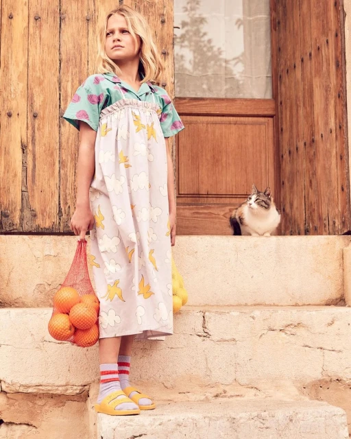 A young girl in a patterned dress stands on stone steps holding a mesh bag of oranges, with a cat visible on the steps in the background.