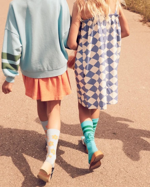 Two young girls walk on a paved surface wearing a blue sweatshirt, orange skirt, blue-and-white dress, patterned socks, and sandals.