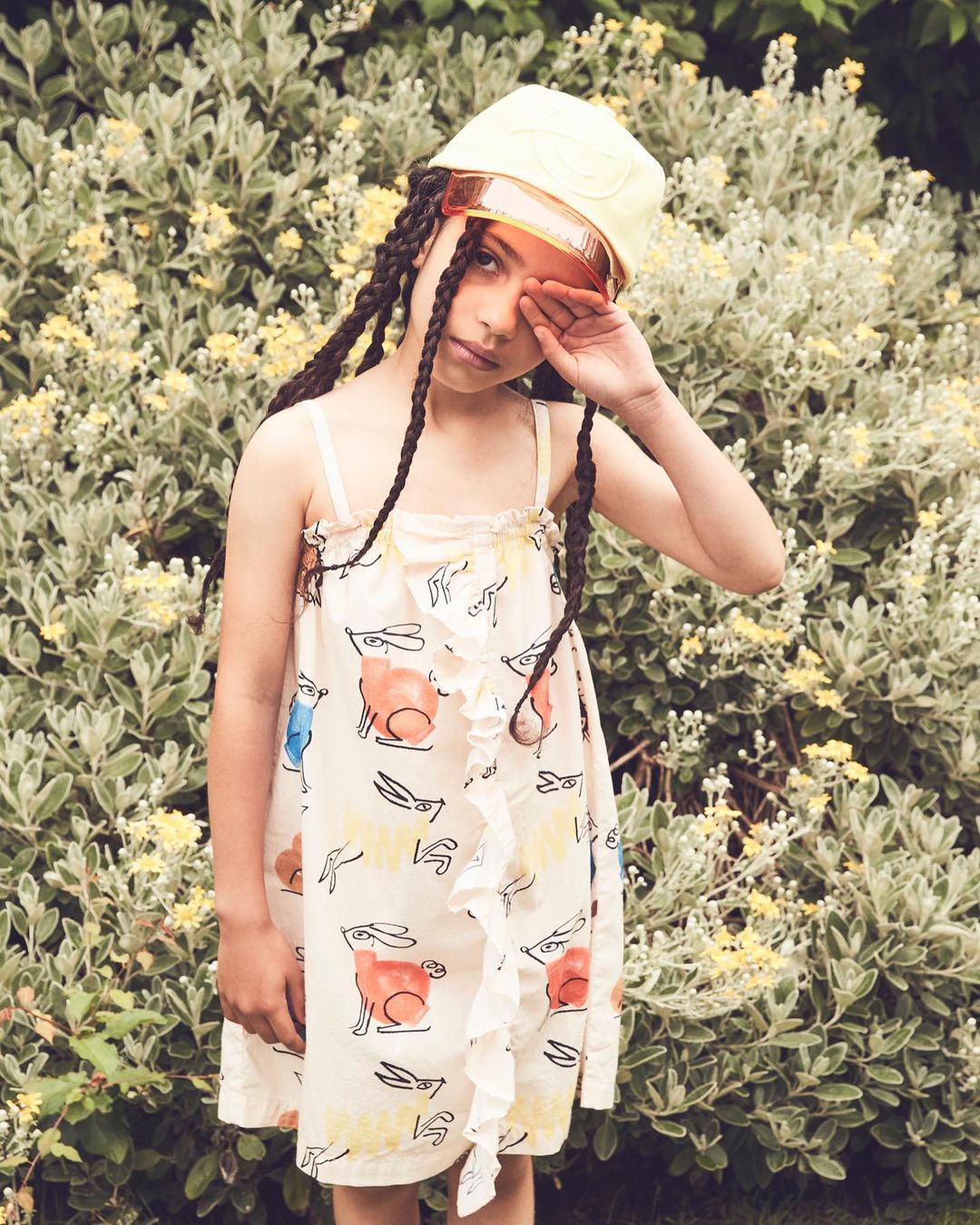 Young girl in printed dress and visor poses in front of flowering green bush in natural light. Fashion portrait of a child model.