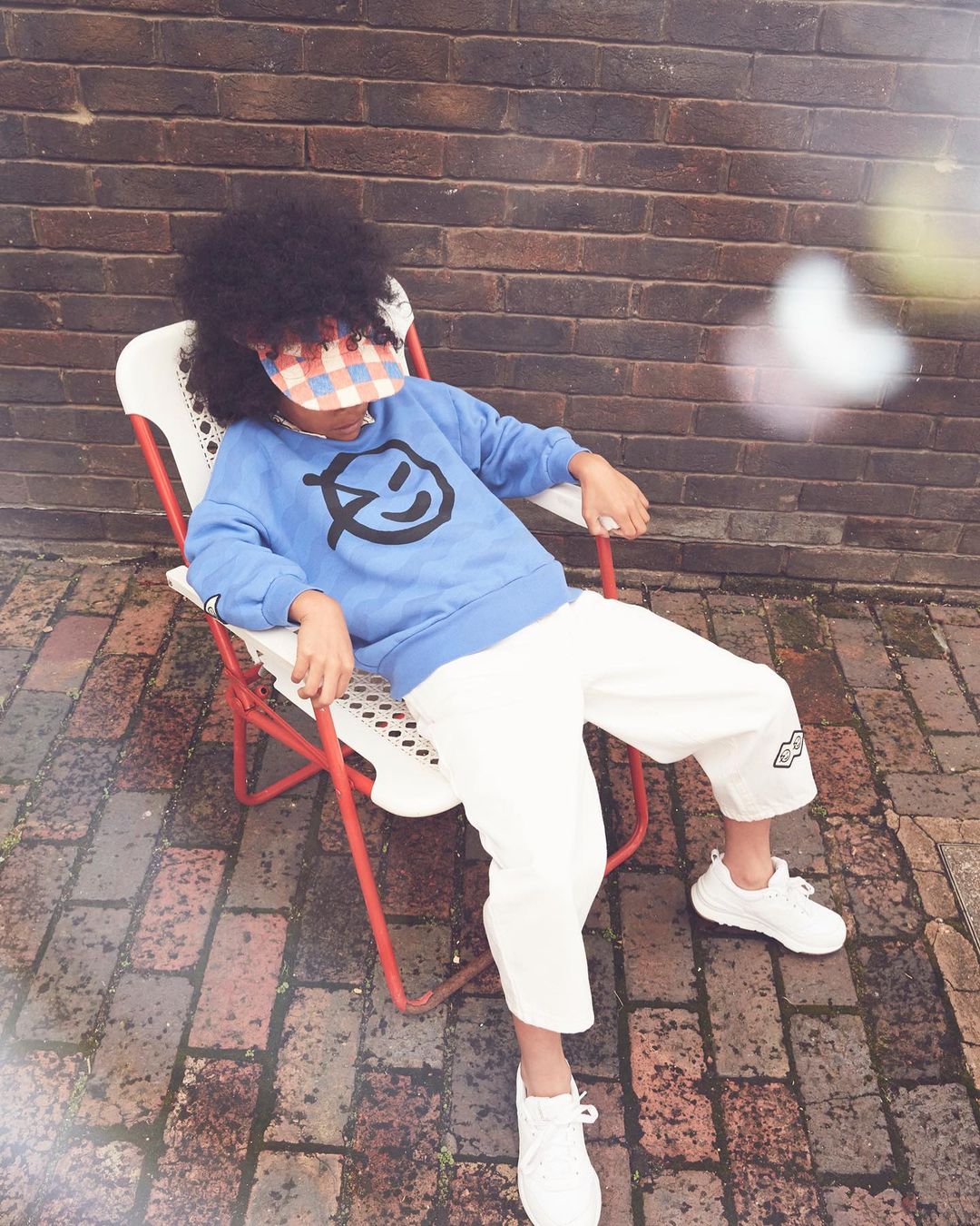 Child with afro sits in an outdoor chair wearing a graphic sweatshirt, white pants, white sneakers, and visor against a brick wall.