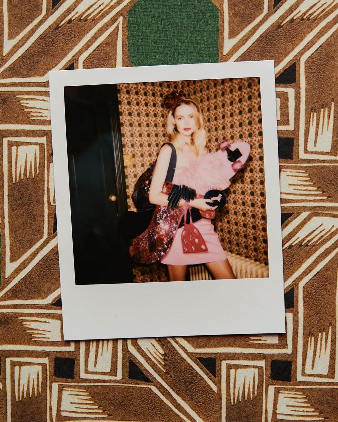 Photo booth style portrait of a blond woman in a pink dress holding feather boa, posing with a sequined bag in a patterned room.