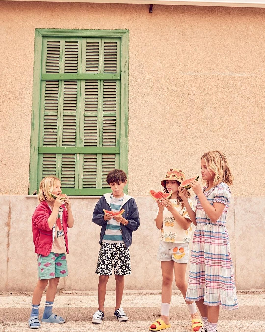 Four children stand outdoors in front of a building with a green shuttered window, eating watermelon slices, wearing shorts and summer clothing.