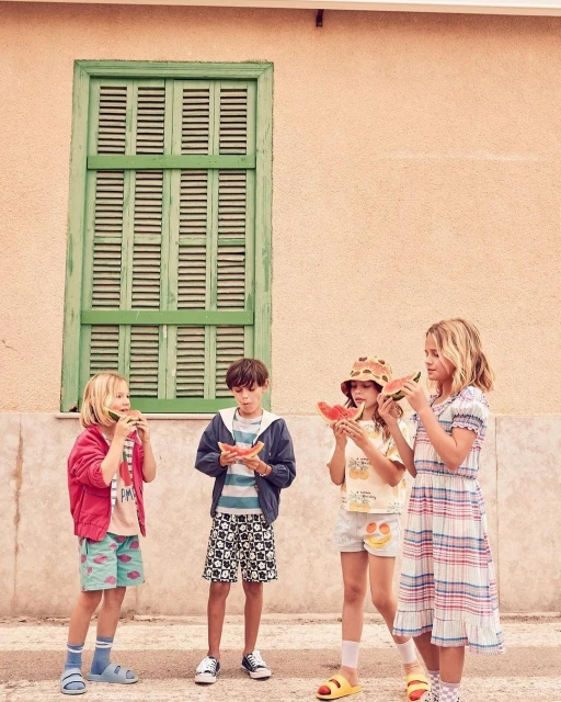 Four children stand outdoors in front of a building with a green shuttered window, eating watermelon slices, wearing shorts and summer clothing.