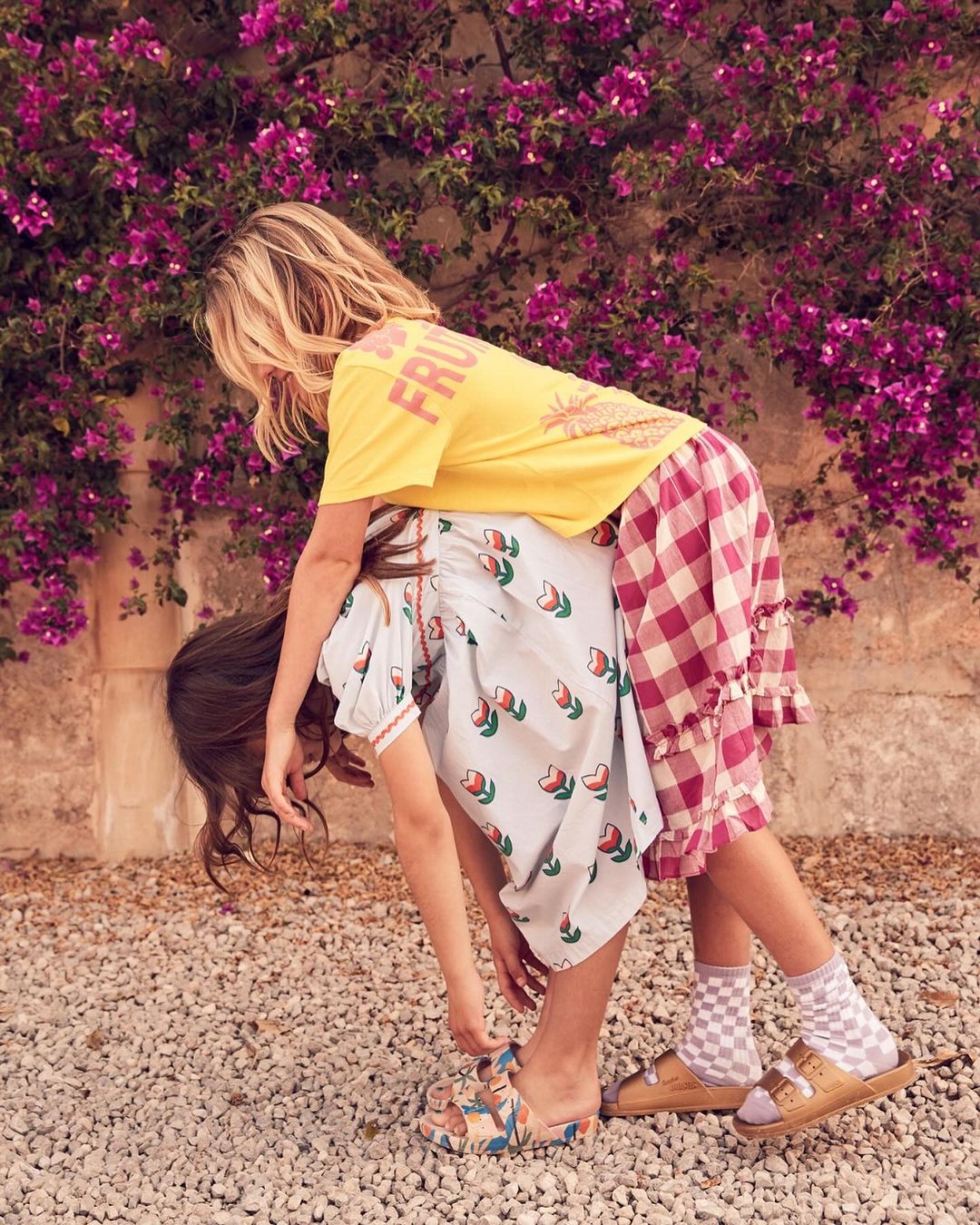 Two young girls in floral and checkered dresses playfully bending over outdoors, showcasing children's fashion with a vibrant, fun aesthetic.