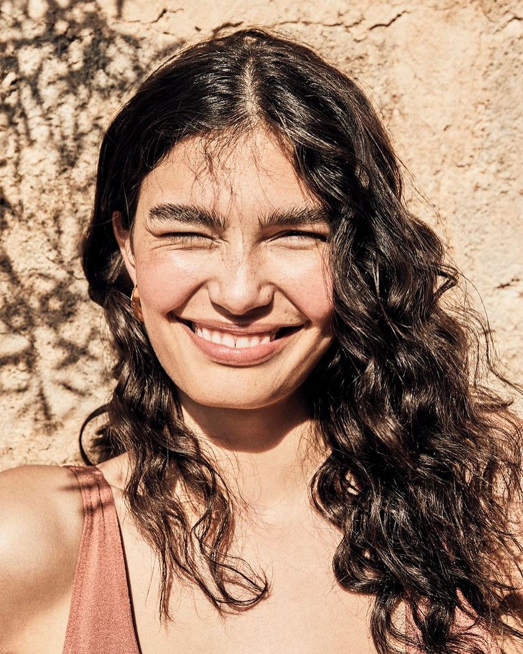 Close-up portrait of an adult woman with curly brown hair smiling with her eyes closed in front of a textured wall with shadows.