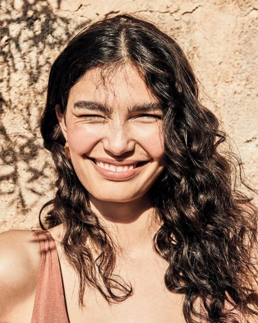 Close-up portrait of an adult woman with curly brown hair smiling with her eyes closed in front of a textured wall with shadows.