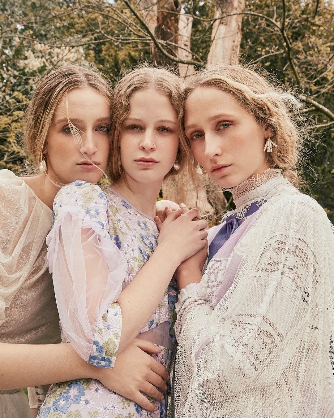 Three fair-skinned young women pose closely together in an outdoor setting, wearing floral dresses and vintage-inspired lace garments.