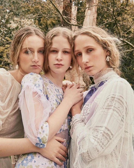 Three fair-skinned young women pose closely together in an outdoor setting, wearing floral dresses and vintage-inspired lace garments.