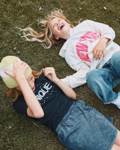 Two laughing girls lie on the grass in casual clothing. One wears a hat and a t-shirt, while the other wears a hooded sweatshirt.