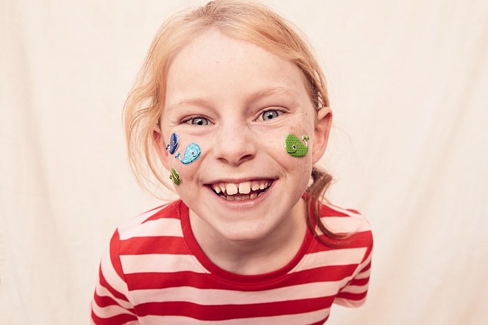Happy young girl with blond hair and stickers on her face, wearing a red and white striped shirt. Smiling, close-up shot.