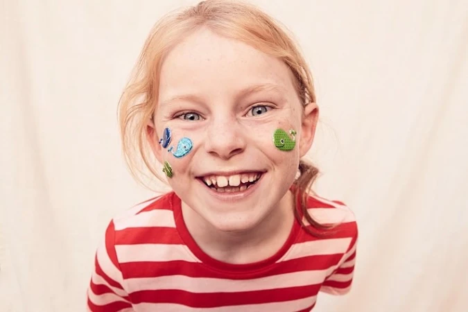 Happy young girl with blond hair and stickers on her face, wearing a red and white striped shirt. Smiling, close-up shot.