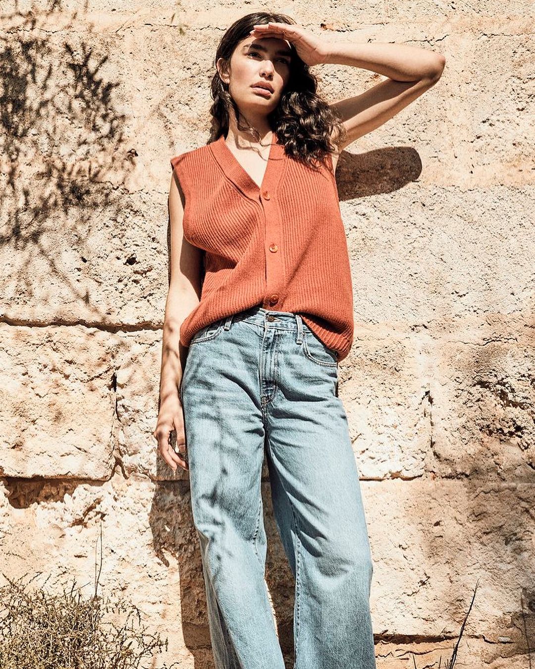 A brunette woman shields her eyes from the sun near a textured rock wall, wearing a ribbed orange vest and wide-leg jeans in bright daylight.