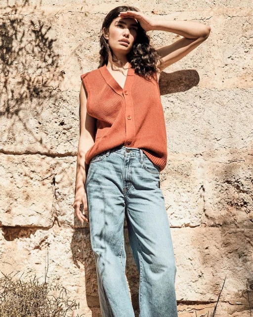 A brunette woman shields her eyes from the sun near a textured rock wall, wearing a ribbed orange vest and wide-leg jeans in bright daylight.