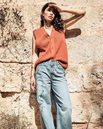 A brunette woman shields her eyes from the sun near a textured rock wall, wearing a ribbed orange vest and wide-leg jeans in bright daylight.