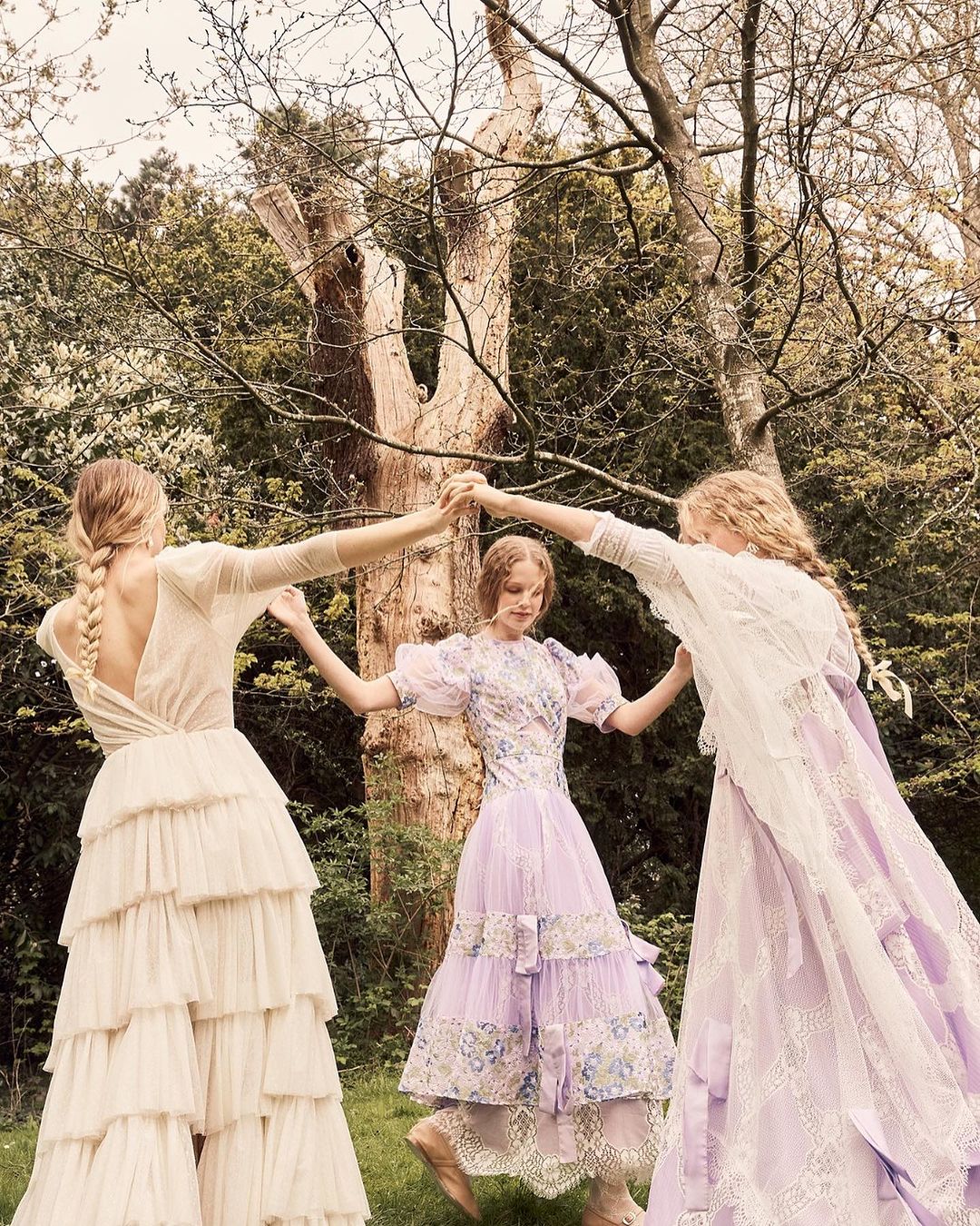 Three women in ornate dresses hold hands in a circle surrounded by greenery. The dresses are light colors with ruffles and lace.