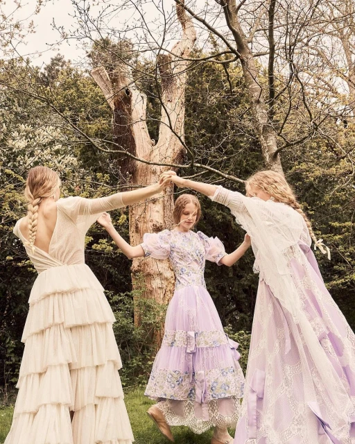 Three women in ornate dresses hold hands in a circle surrounded by greenery. The dresses are light colors with ruffles and lace.
