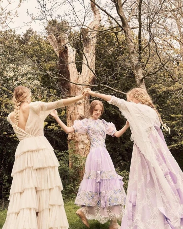 Three women in ornate dresses hold hands in a circle surrounded by greenery. The dresses are light colors with ruffles and lace.