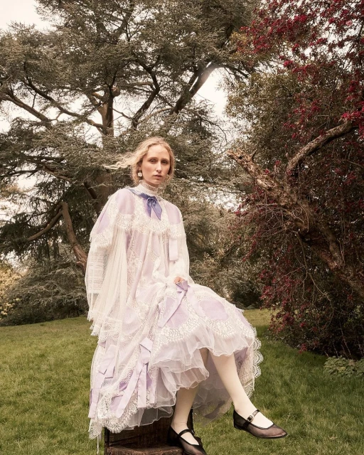 A young fair-skinned woman with long blonde hair sits on a tree stump in a garden wearing a vintage-style dress with lace and bows.