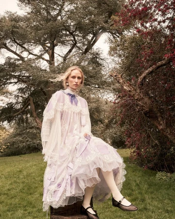 A young fair-skinned woman with long blonde hair sits on a tree stump in a garden wearing a vintage-style dress with lace and bows.
