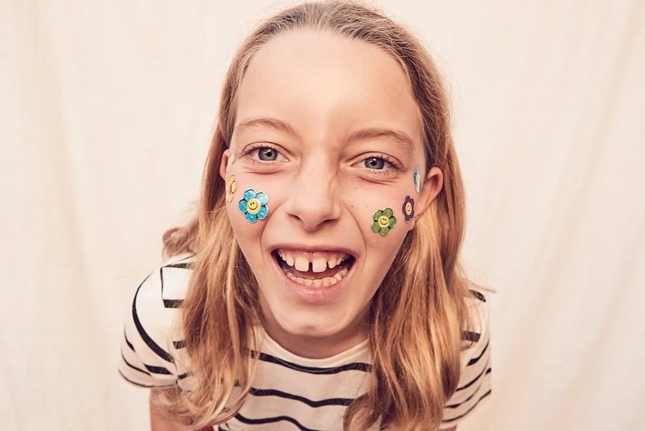 Smiling child with flower face stickers, wearing a striped shirt against a light backdrop, captured in a playful, wide-angle, close-up shot.