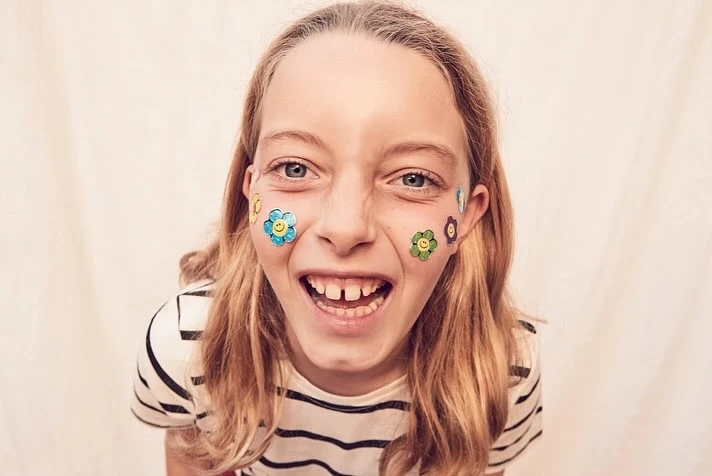Smiling child with flower face stickers, wearing a striped shirt against a light backdrop, captured in a playful, wide-angle, close-up shot.