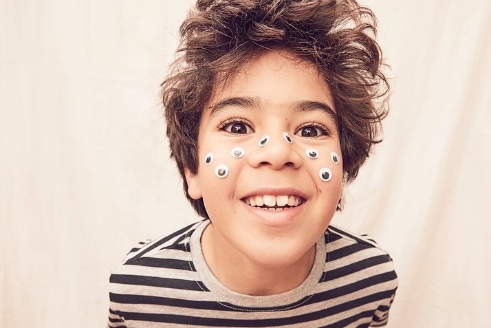 Smiling child with googly eyes on their face, wearing a striped shirt, captured in a close-up shot with a soft, neutral background.