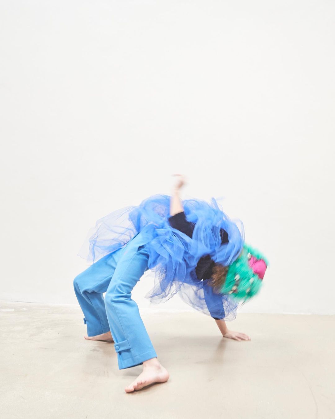 Artistic shot of a person in a blue outfit and a colourful, patterned hat doing a backbend against a light-colored wall and floor.