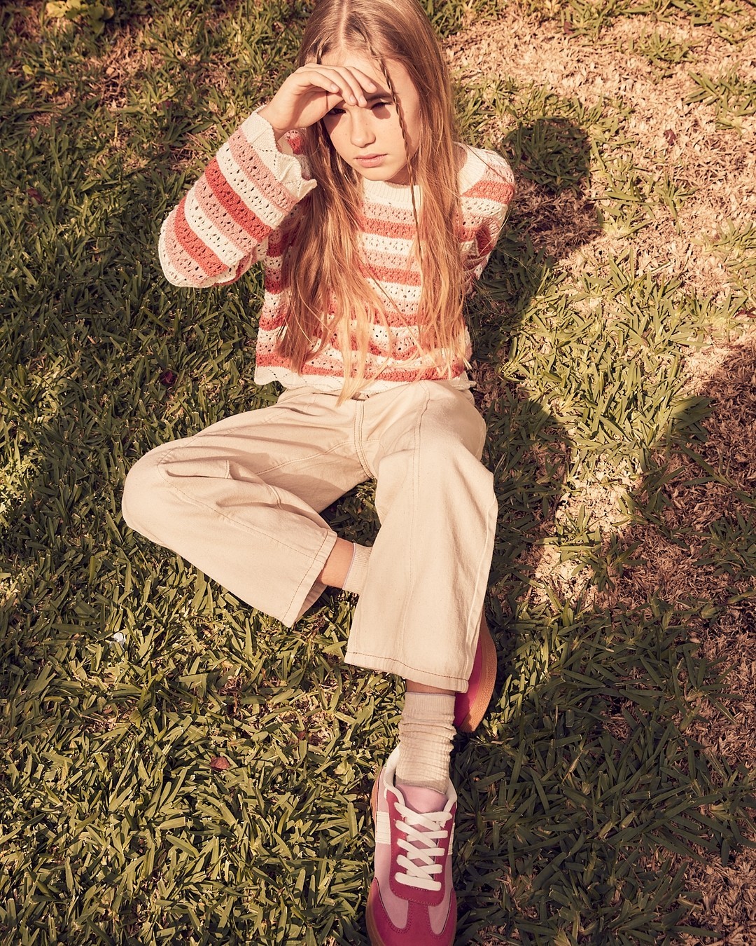 Young model sitting on grass wearing striped knit top, light trousers, and pink sneakers, photographed by Annie Bundfuss