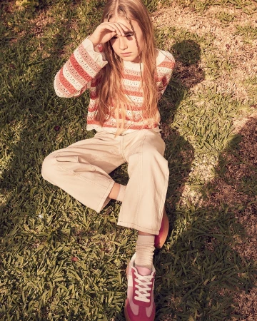 Young model sitting on grass wearing striped knit top, light trousers, and pink sneakers, photographed by Annie Bundfuss