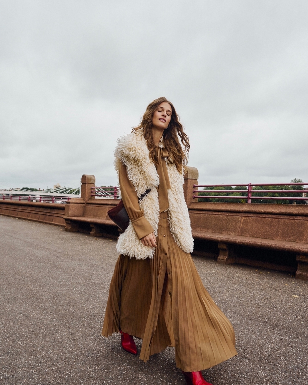 Fashionable adult female posing on an urban bridge wearing a sleeveless fur jacket, tan dress and red boots with an overcast sky.