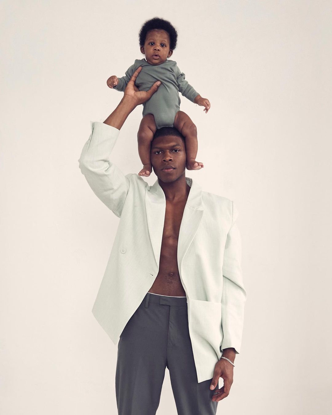Studio shot of an adult man holding a baby overhead against a neutral background. The man is wearing an open suit jacket with no shirt.