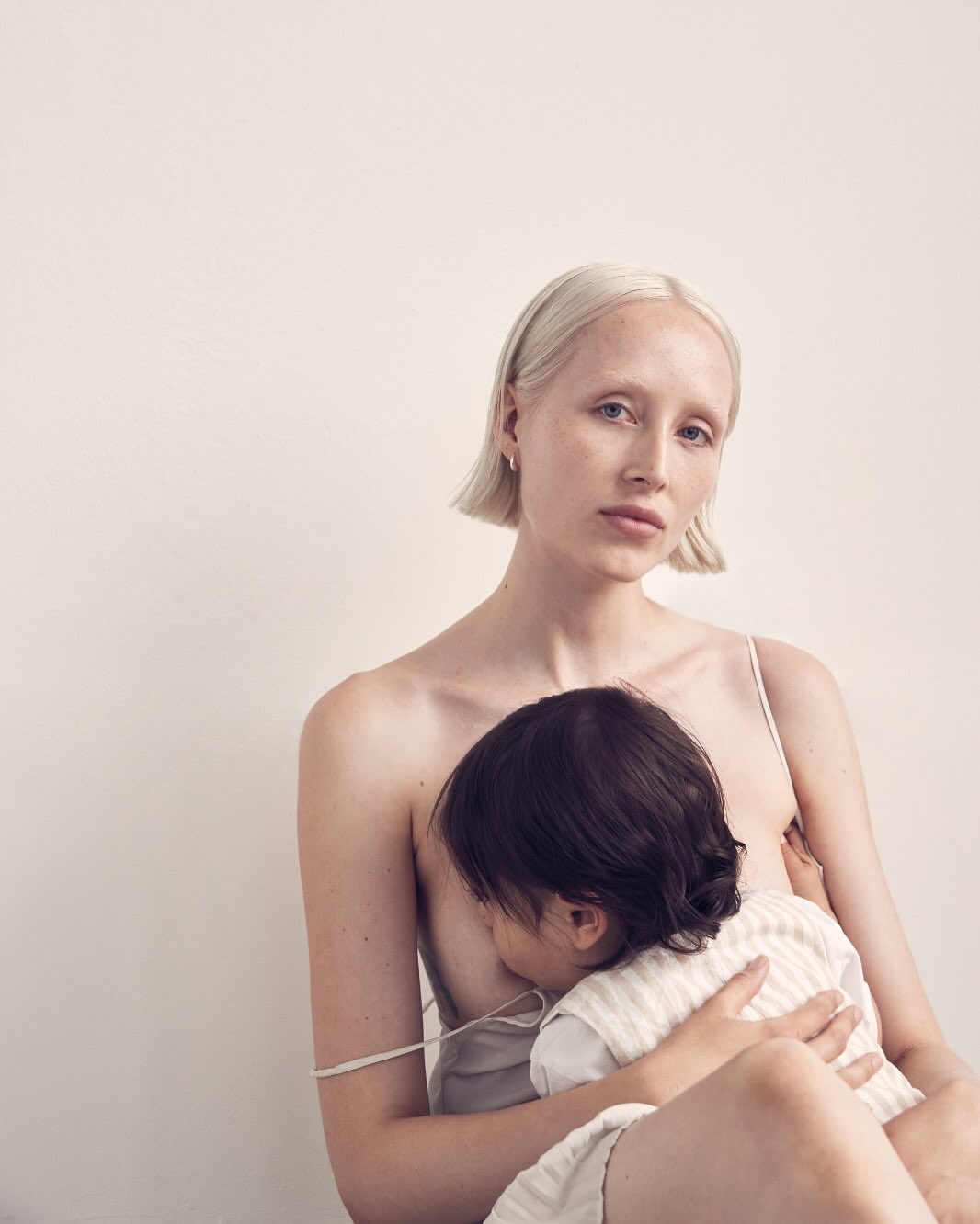 Blonde woman breastfeeding child indoors against a white wall in soft light, creating a nurturing and intimate maternal moment.