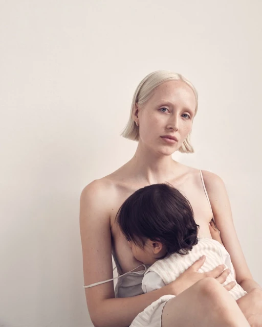 Blonde woman breastfeeding child indoors against a white wall in soft light, creating a nurturing and intimate maternal moment.