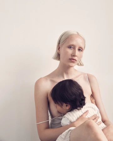 Blonde woman breastfeeding child indoors against a white wall in soft light, creating a nurturing and intimate maternal moment.