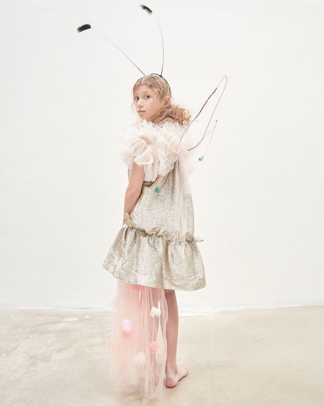 A young girl in a fairy costume with a gold dress, wings, and a feathered headband, stands in front of a bright white background, looking at the viewer.