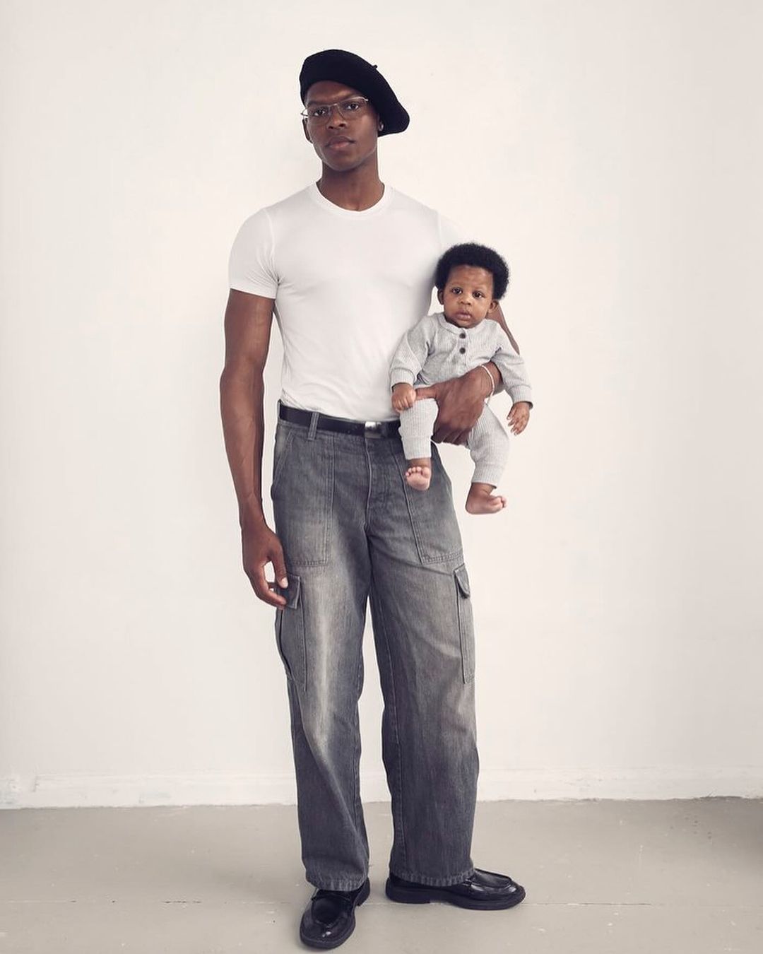 An adult male wearing a beret and wide-legged jeans holding a baby in a gray jumpsuit, both looking at the camera against a white backdrop.