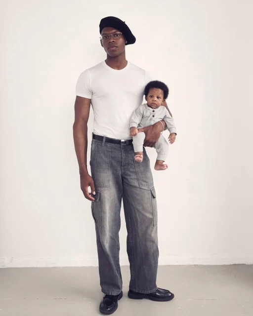 An adult male wearing a beret and wide-legged jeans holding a baby in a gray jumpsuit, both looking at the camera against a white backdrop.