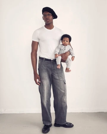 An adult male wearing a beret and wide-legged jeans holding a baby in a gray jumpsuit, both looking at the camera against a white backdrop.