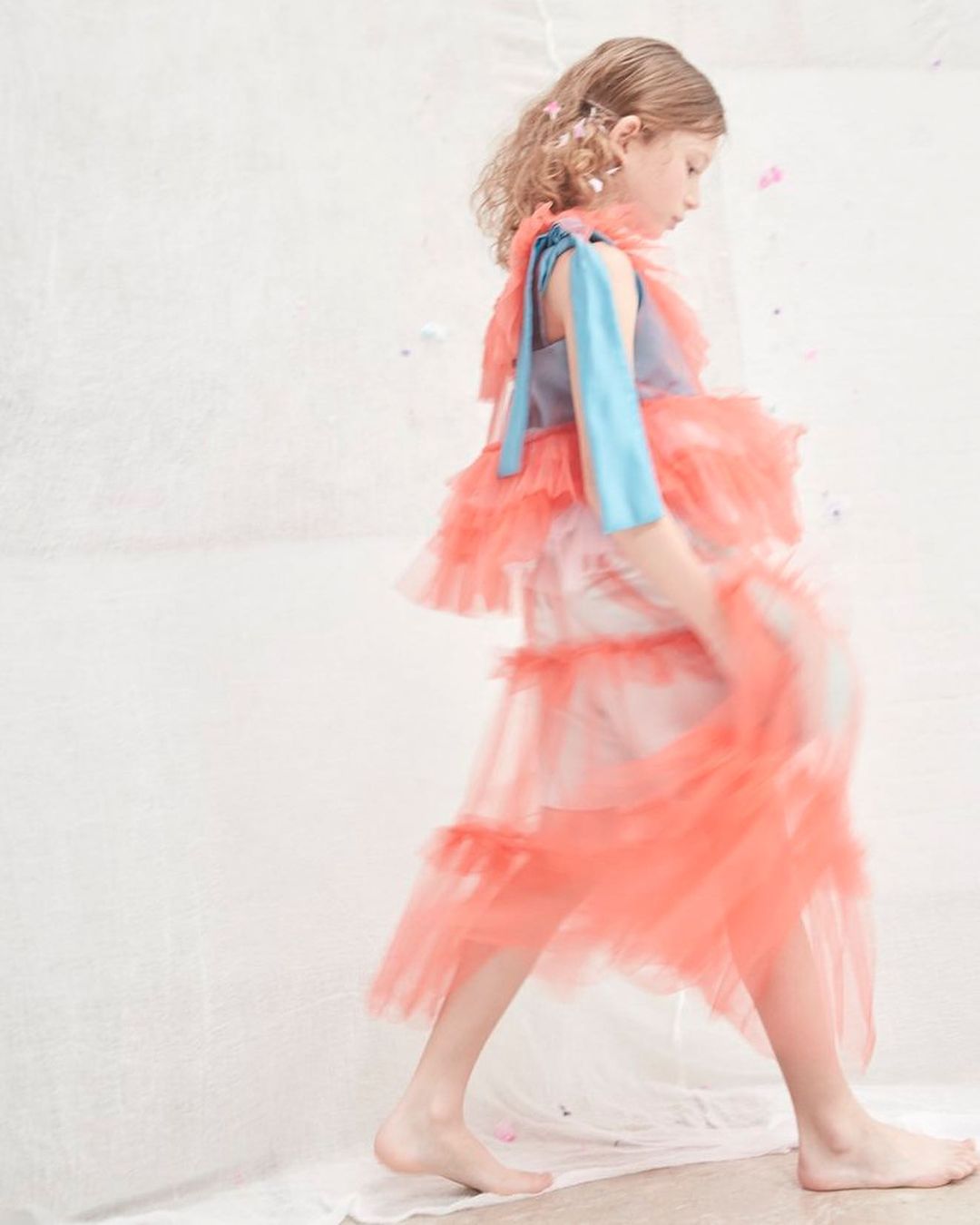 A young girl in a layered coral dress poses in a light-filled studio with a textured white backdrop, captured in a flowing, motion-blurred style.