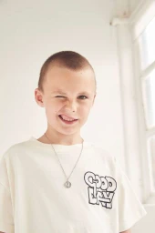 Close-up portrait of a child with a buzz cut winking and smiling, wearing an off-white t-shirt with graphic text.