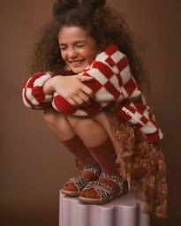 Young child with curly hair crouched and laughing, wearing a red and white checkered fleece jacket, patterned skirt, and sandals.