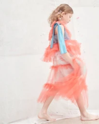 Young girl walking barefoot in a layered pink tulle dress with blue straps against a white seamless backdrop